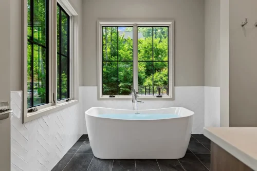 Primary bathroom with a freestanding soaking tub, large windows overlooking the woods, white tile walls, and dark tile flooring.