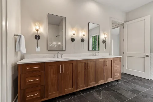 Primary bathroom with a long double vanity, warm wood cabinetry, stone countertop, wall sconces, and dark tile flooring.