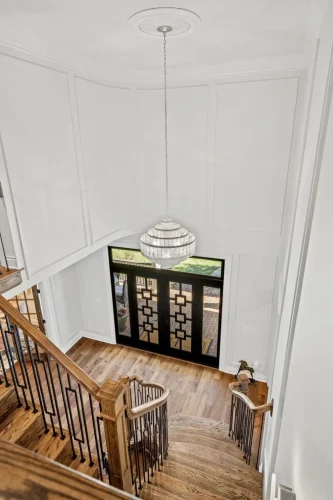 Two-story foyer featuring a curved wooden staircase, black iron railings, white paneled walls, a crystal chandelier, and decorative glass front doors.