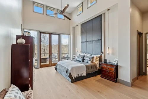 Spacious bedroom with a tall ceiling, oversized upholstered headboard, wood flooring, and glass doors opening to a balcony.