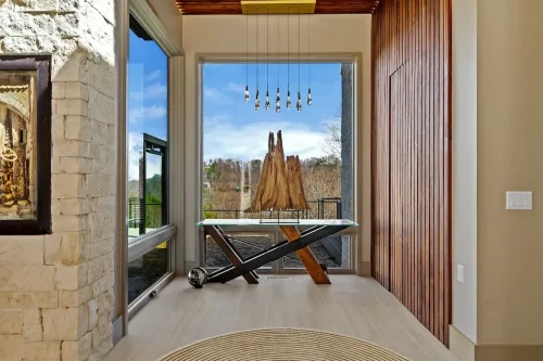 Sculptural wood display on a glass table framed by large windows, a wood accent wall, and pendant lighting.