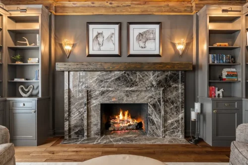 Marble fireplace with a rustic wood mantel, dark gray built-in bookshelves, decorative wall sconces, and framed horse artwork.