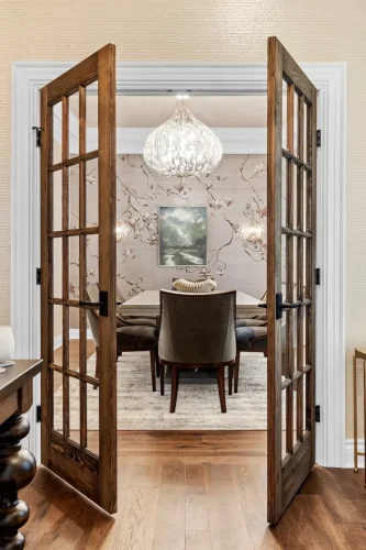 Wood-framed glass French doors opening into a formal dining room with floral wallpaper, upholstered chairs, and a decorative chandelier.