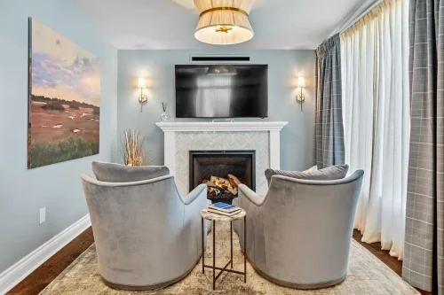 Sitting area with two upholstered chairs, a fireplace with a herringbone tile surround, wall-mounted television, and large windows with curtains.