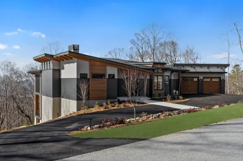 Contemporary mountain home with wood, stone, and dark siding accents, featuring a three-car garage and landscaped driveway on a sloping lot.