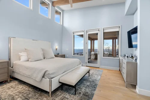 Spacious bedroom with tall ceilings, wood beams, and soft blue walls, featuring a modern bed and glass doors opening to a deck with mountain views