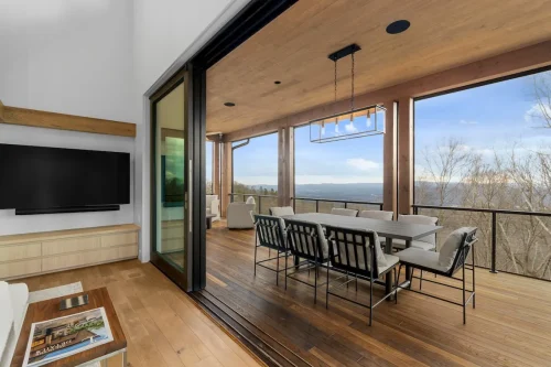Covered deck with long dining table, cushioned chairs, and panoramic views of the Blue Ridge Mountains, connected to the living room through sliding glass doors.