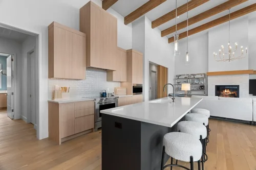 Open-concept kitchen with white quartz island, pendant lighting, and wood cabinetry, leading into a bright living room with fireplace and vaulted ceilings with wood beams.