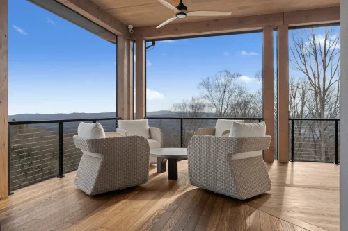 Cozy seating area on a covered deck with woven lounge chairs around a round coffee table, overlooking wooded mountain views.