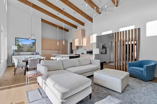 Expansive great room with vaulted ceilings, exposed beams, modern chandelier, sectional sofa, and open flow to kitchen and dining spaces.