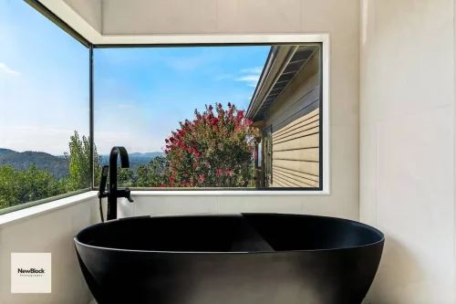 Black bathtub beside large window with red flowering tree outside.