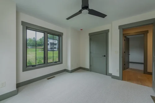 Bedroom with neutral carpet, green trim, ceiling fan, and large window overlooking the neighborhood.