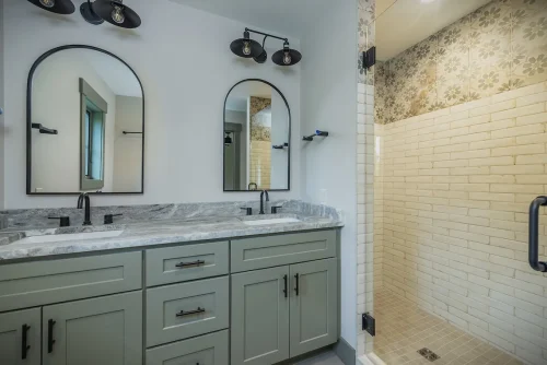 Primary bathroom with double vanity, arched mirrors, green cabinetry, and walk-in shower with decorative tile accent.
