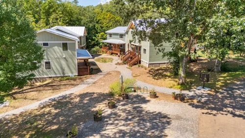 Aerial view of a residential community with green-sided homes, metal roofs, wooden porches, gravel pathways, and surrounding trees.