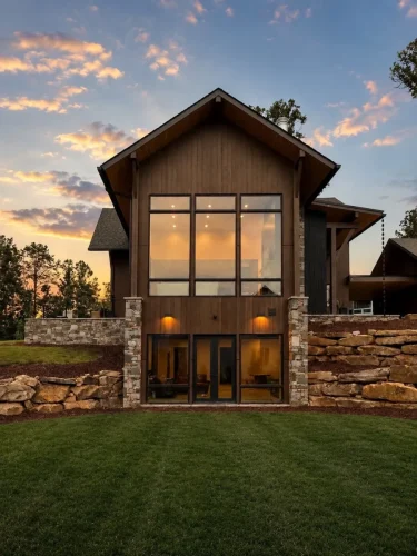 Exterior view at sunset featuring tall black-framed windows, wood siding, stone accents, and a walkout lower level.