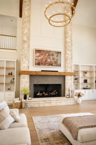 Bright two-story great room featuring a floor-to-ceiling stone fireplace, built-in shelving, exposed wood beams, and a large circular chandelier