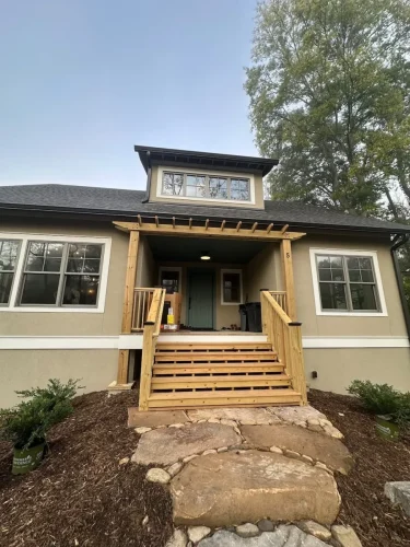 Front view of a home with light-colored siding, a dormer window, covered entry, natural wood porch and steps, and stone walkway.