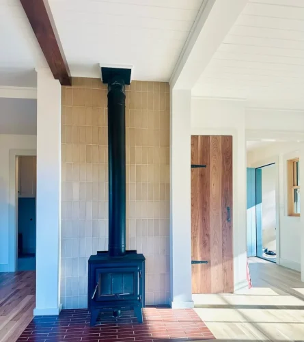 Black freestanding wood stove against a beige tile accent wall, featuring a brick hearth, white paneled ceiling, and exposed wood beam.