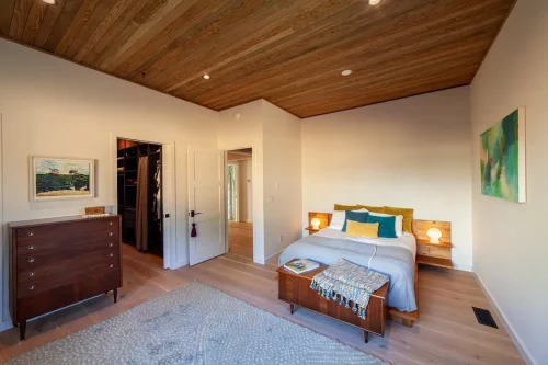 Bedroom with a wood ceiling, light wood floors, colorful pillows, a wooden chest at the foot of the bed, and framed artwork.
