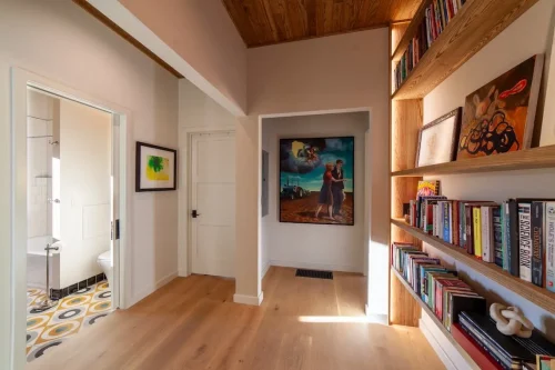 Hallway with built-in wood bookshelves, colorful artwork, and an open doorway revealing patterned bathroom flooring.