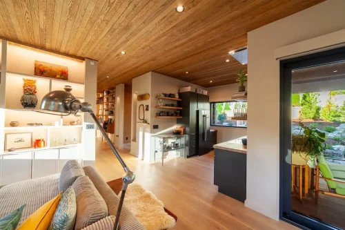 Open living area with a wood ceiling, illuminated display shelves, a striped sofa, and views into the kitchen and outdoor seating area.