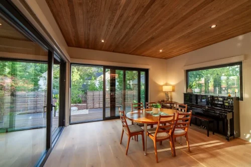 Dining room with a round wood table, an upright piano, a wood ceiling, and large glass doors opening onto a deck.