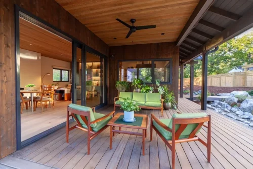 Covered wood deck with green-cushioned seating, potted plants, a ceiling fan, and sliding glass doors leading to the dining room.