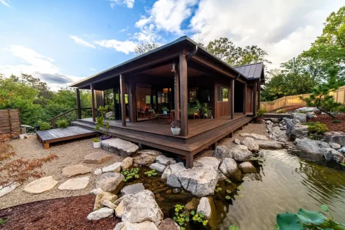 Wood-sided home with a covered deck overlooking a landscaped pond bordered by large rocks and garden plants.