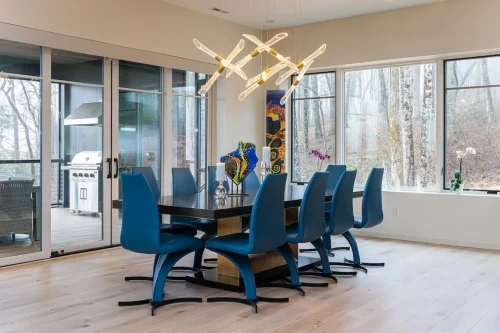 Dining area with a dark wood table, blue upholstered chairs, sculptural chandelier, and large windows overlooking the wooded landscape.