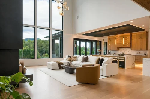 Bright open-concept living room with soaring windows, cozy white sectional sofa, modern armchair, and views into the sleek kitchen and mountain scenery beyond.