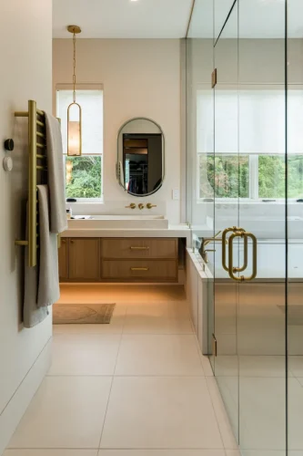 Bright bathroom with glass shower, wood vanity, oval mirror, and brass fixtures.