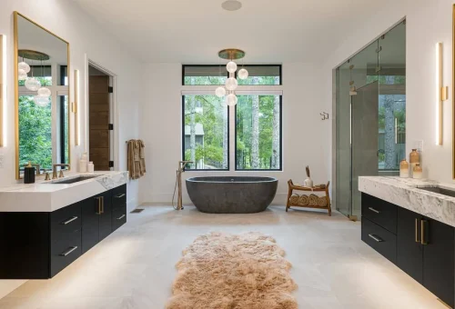 Spa-like bathroom with dual vanities, marble countertops, a freestanding stone soaking tub beneath a modern chandelier, and glass shower with views of the forest outside.