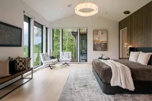 Sophisticated bedroom with dark accent wall, plush bed with layered linens, sitting area with modern chairs, large windows overlooking greenery, and elegant lighting fixtures.
