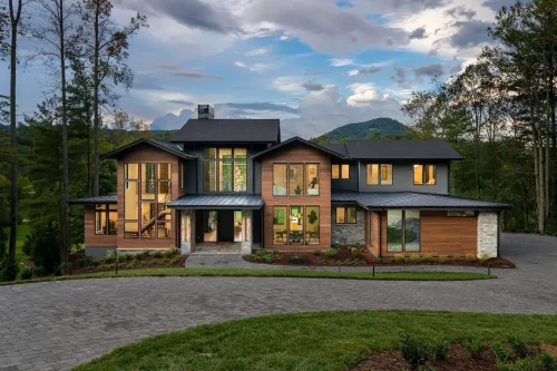 Striking modern home at dusk with warm interior lights glowing through expansive windows, wood and stone facade, and mountain backdrop beneath a dramatic sky.