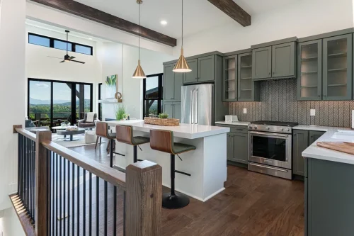 Open kitchen with green cabinetry, a large white island, pendant lighting, exposed ceiling beams, and an adjacent living area.
