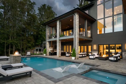 Resort-style outdoor living space with a sleek pool, fire features, lounge chairs, and covered patios, backed by tall trees and dramatic evening lighting.