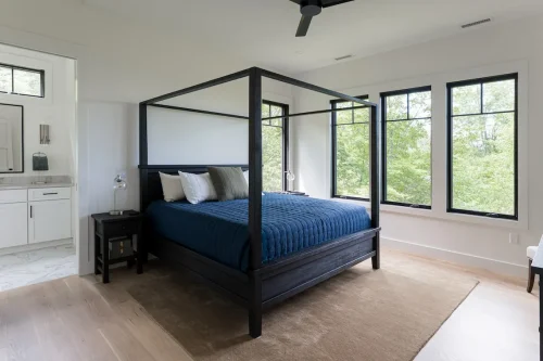 Bedroom with black canopy bed, blue quilt, and large windows overlooking trees.
