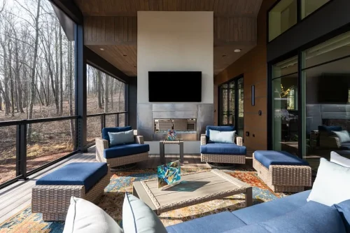 Screened outdoor living area with blue cushioned seating, a fireplace, mounted television, and wood-paneled ceiling overlooking the woods.