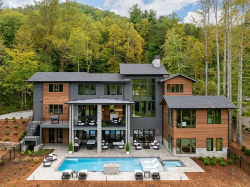 Expansive modern mountain home with wood and dark siding, soaring windows, and multiple balconies overlooking a resort-style backyard with a swimming pool, fire feature, and lounge seating surrounded by forest.