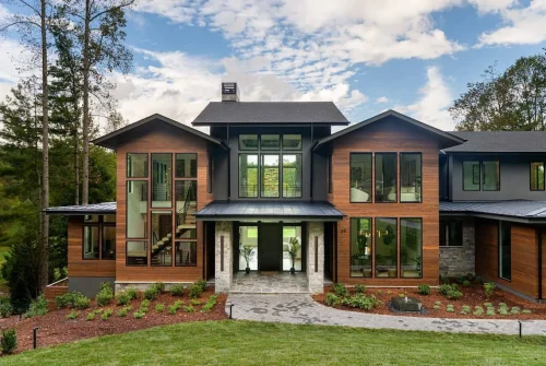 Modern mountain home with wood siding, large glass windows, and stone accents, framed by tall trees and landscaped front yard under a bright blue sky.