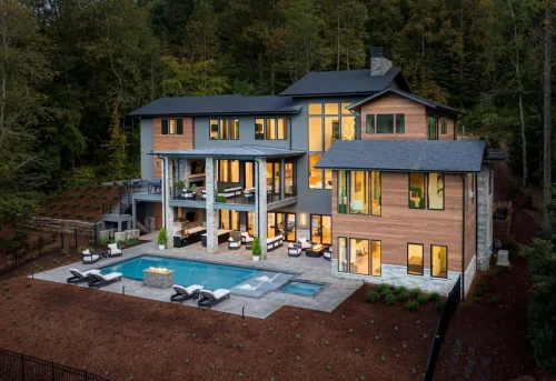 Luxury home at dusk with warm interior lighting glowing through large glass windows, covered patios, and a sleek pool terrace featuring loungers, firepit, and spa, all set against wooded hills.