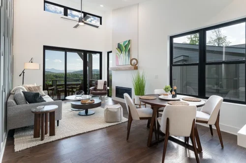 Open living and dining space with a fireplace, tall windows, dark wood flooring, and doors opening to a mountain-view deck.