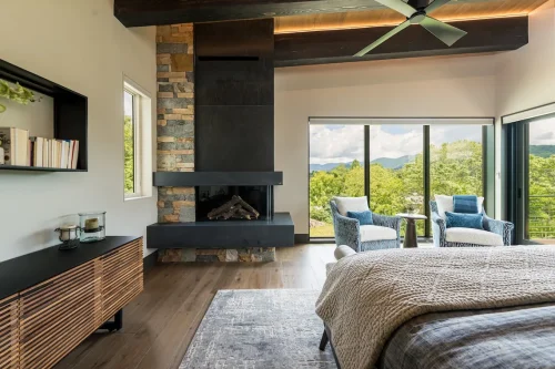 Modern bedroom with stone fireplace, wood floors, large windows, and mountain views.