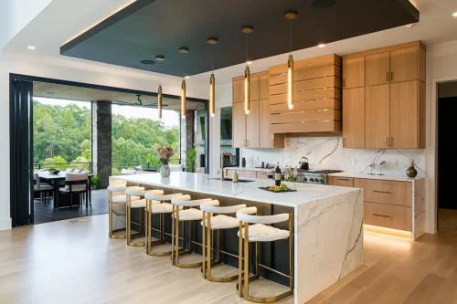 Sleek open kitchen featuring a marble waterfall island with gold barstools, light wood cabinetry, and pendant lighting, opening to a covered patio with mountain views.