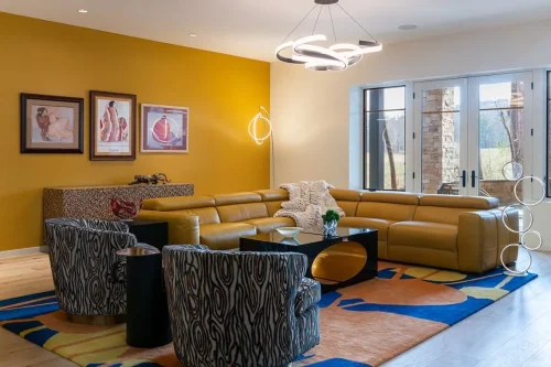 Colorful living area featuring a mustard yellow accent wall, leather sectional sofa, patterned chairs, and modern chandelier.