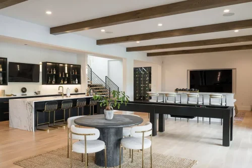 Spacious game and bar area with exposed wood beams, marble bar counter, modern seating, shuffleboard table, and a wall-mounted TV beside a glass wine display.