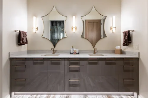 Double vanity with glossy gray cabinetry, marble countertops, decorative mirrors, and gold-accented lighting.