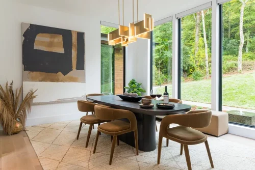 Contemporary dining room with floor-to-ceiling windows, a dark modern table with leather chairs, abstract wall art, and statement gold pendant lighting overlooking the backyard.