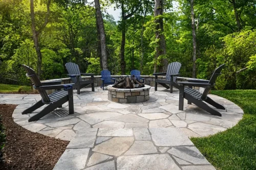 Stone patio with Adirondack chairs surrounding a circular fire pit in a wooded setting.