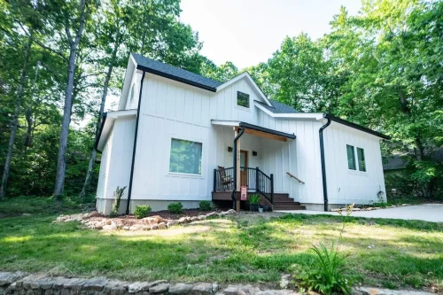 Modern farmhouse-style home with white board-and-batten siding, black trim, and a welcoming covered porch framed by trees.